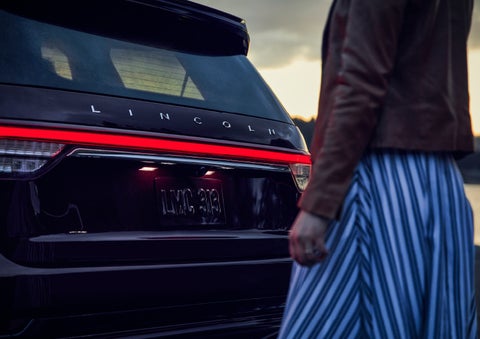 A person is shown near the rear of a 2024 Lincoln Aviator® SUV as the Lincoln Embrace illuminates the rear lights | Mike Reichenbach Lincoln in Florence SC